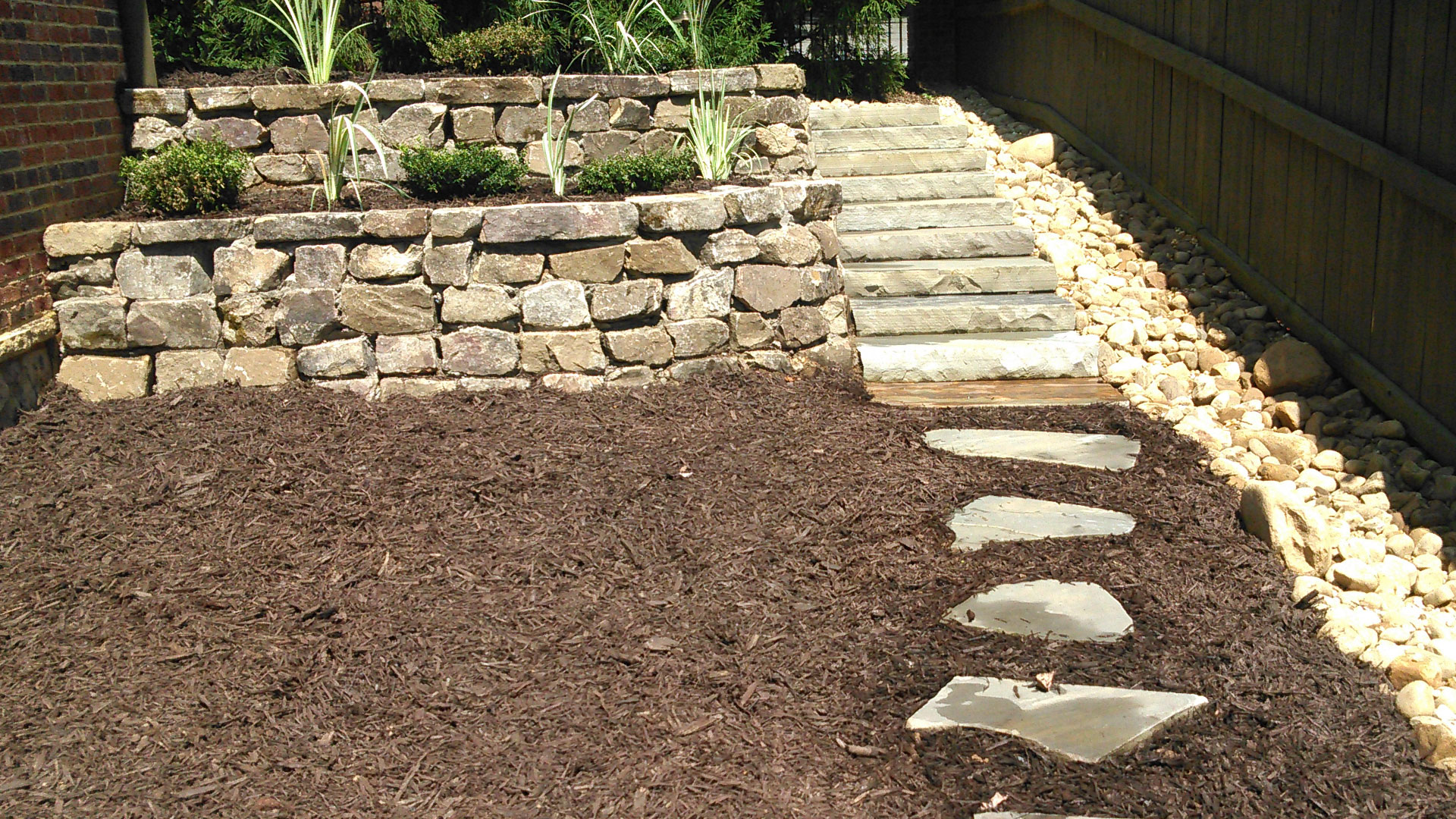 A small rock retaining wall with steps, small plants, and mulch